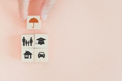 senior's hand hold wooden cube block screen  umbrella icon above family, house, graduation hat and car symbol for insurance concept