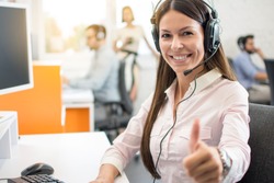 Portrait of beautiful young woman wearing headset showing thumbs up gesture. Customer support service agent woman working in call center and showing success sign with a hand.