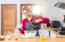 Asian girl using the blender, blend dough and other ingredients for cooking the chocolate cake with happy feeling in the kitchen room. 