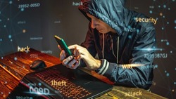 A hacker in a hood with a phone is typing on a laptop keyboard in a dark room. The concept of cybercrime fraud and identity theft