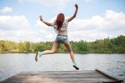 Photo from back of young jumping woman on wooden bridge by river