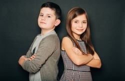 Studio portrait of two cute kids, standing back to back, arms crossed, posing on black background