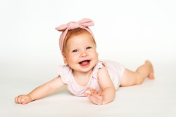 Portrait of adorable red-haired baby lying on belly, white background, wearing pink body and headband