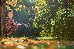 Young woman sitting under an autumn tree leaning on the tree trunk enjoying and relaxing.
