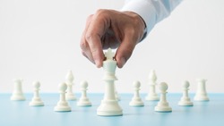 Closeup of male hand holding white chess figure of king positioned in front of the other figures on a blue desk in a conceptual image.