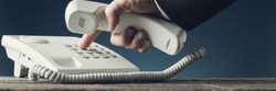 Wide view image of businessman dialing telephone number on white landline phone while holding a handset. Over navy blue background.