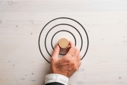 Hand of a businessman placing blank wooden cut circle in the middle of a target drawn on white textured wooden background. 
