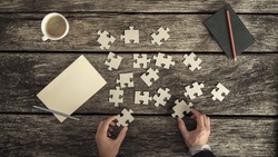 Retro style image of male hands in business suit trying to find a solution to a problem by arranging and matching puzzle pieces on a textured rustic wooden desk, top view.