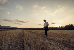 Retro image of businessman in elegant suit holding his jacket standing in mown wheat field under a majestic evening sky with a setting sun.