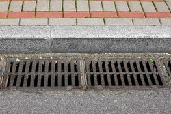 catch basin grate of the lattice of the drain system for drainage of rainwater from the road surface with a tarmac road at the curb with a stone brick tile sidewalk close-up, nobody.