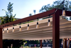 textile awning in the backyard wooden frame gazebo with a garland of strings of retro edison light bulbs of lamps lighting glowing with warm light on sunny summer day close-up, nobody.
