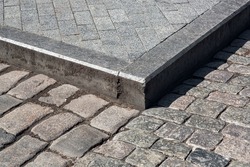 corner of a pedestrian sidewalk made of stone rectangular brick tiles and granite curb dirty in mud near a road paved with rough cobblestone close-up on sunny day, nobody.