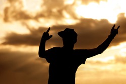 A man in a cap and raised arms takes pleasure at an outdoor music festival. Black silhouette on sunset