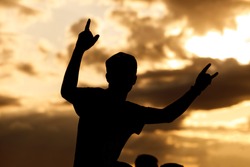 A man in a cap and raised arms takes pleasure at an outdoor music festival. Black silhouette on sunset