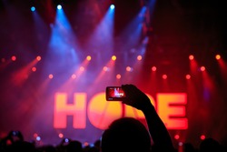 Hand with a smartphone records live music festival, Taking photo of rock concert stage, live concert, music festival. The word - hope - on stage, red light