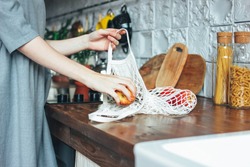 Young woman in the grey dress pulls apples out of knitted rag bag string bag shopper in the kitchen, zero waste, slow life