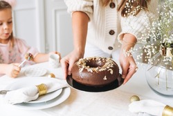 Young woman mother in white holding in hands Large round chocolate almond cake on table with New Year serving, christmas white scandinavian festive table, family event