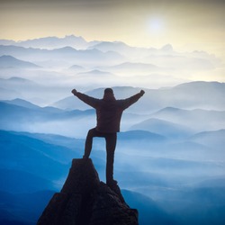 Hiker who conquered the top of mountain standing on a cliffs edge with raised hands and enjoy sunrise in a misty valley.