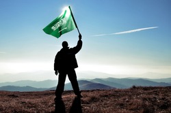 successful silhouette man winner waving Saudi Arabia flag on top of the mountain peak