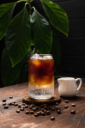 Cold espresso tonic. A highball glass filled with ice cubes, tonic soda water, coffee beans around, milk jug, wooden background