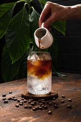 Espresso tonic in the making. Adding coffee into a highball glass filled with ice cubes and tonic soda water, coffee beans, milk jug, wooden background