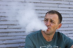 A middle-aged man sitting on a background of gray brick wall and releases the smoke out of her mouth/ Portrait of a man who smokes/ Man blows smoke