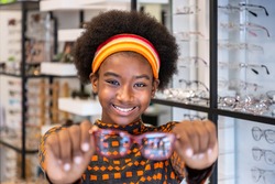 Portrait of Young African woman girl with Afro hair style choosing glasses in optical shop, holding eyeglasses in hand, smiling, looking at camera. Eyecare concept.