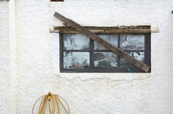 Closeup of the white wall of a house  with an old barricaded window with dirty panes and a yellow water hose dangling from a tap. 