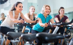 Cheerful smiling different age females cycling on exercise bikes at fitness club 