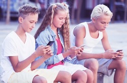 Modern positive kids spending time together outdoors using mobile gadgets