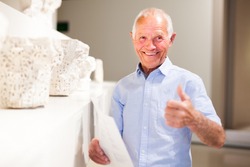 Smiling senior man posing in art exhibition and showing thumbs up