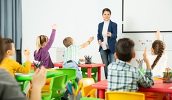 Smiling young woman teacher examining pupils at lesson in elementary school. Children raising hands to answer
