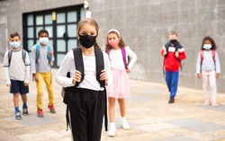 Confident teen girl in protective mask walking outside school building on spring day, going to lessons. Concept of necessary precautions in COVID pandemic.