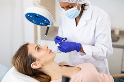 Hispanic woman cosmetician giving face injections of botulinum toxin for young European woman.