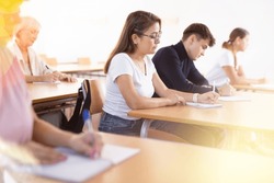 Focused young attractive Hispanic woman listening to lecture and taking notes in classroom with group of adult people. Postgraduate education concept