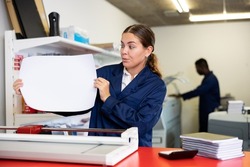 Attentive young female typographer in a blue uniform verifying printed sheet during work in the printer shop
