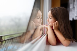 Beautiful little girl smiling and watching out the window. A child looks out the window. Young girl looking from window. Portrait of cheerful kid lies at windowsill.