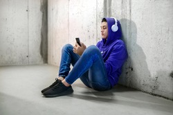 Young teenage boy wearing headphones chatting on his mobile phone as he sits on the concrete floor in a subway or corridor in a blue hoodie and jeans