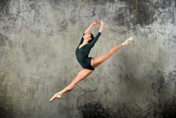 Graceful young ballerina leaping high in the air in a studio portrait against a grey wall with copyspace