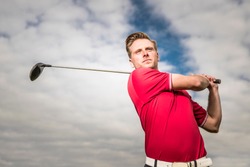 YOUNG GOLF PRO WITH RED SHIRT ILLUSTRATING A SWING ON A GOLFCOURSE WITH A BACKGROUND OF A BLUE SKY WITH WHITE CLOUDS 