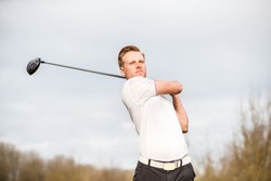 YOUNG GOLF PRO WITH WHITE SHIRT ILLUSTRATING A SWING ON A GOLFCOURSE WITH A BACKGROUND OF A LIGHT SKY WITH WHITE CLOUDS 