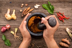 The Women hold pestle with mortar and spice paste of thai popular food red curry  on rustic wooden background. Spices ingredients chilli ,pepper, garlic,galanga lemongrass and Kaffir lime leaves .
