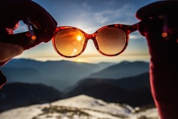 Female hands holding sunglasses through which you can see a beautiful inter sunset in the mountains of blue and orange colors