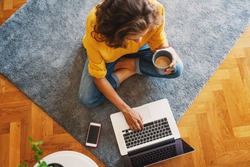 young girl woman in a yellow shirt works on a laptop with a mug of coffee at home in the living room on the carpet, remote work and education