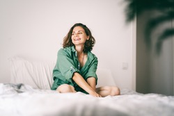 Beautiful charming young curly girl woman in a green shirt sits in bed in the morning and enjoys a new day, shot of a happy smiling face