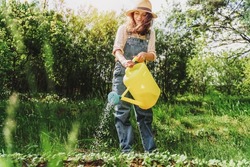 Beautiful happy young woman in a hat watering a bed of yellow watering can in summer garden