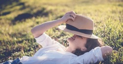 Young happy woman in white blouse lying on green grass covering face with straw hat, summer vacation
