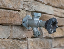 Horizontal close-up shot of an outdoor water faucet attached to a stone wall. The background is partially blurred.