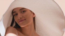 Young gorgeous dark-haired European woman in a big white hat with a smear of sunscreen on her cheek lifts up her shoulder and looks at camera against beige background | Sunscreen concept