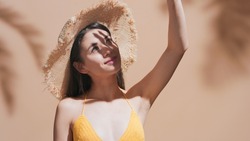 Close-up beauty portrait of young brunette woman in straw hat covers her face from the sun with palm against a beige background | Sunscreen commercial concept
